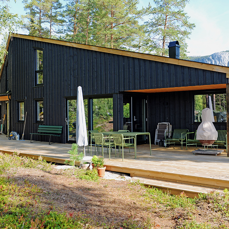 Moderne svart trehytte med terrasse, grønn hagemøbler, hvit parasoll, furutrær.