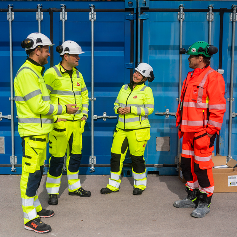 Fire anleggsarbeidere i gule og oransje refleksklær med hjelm foran blå container.