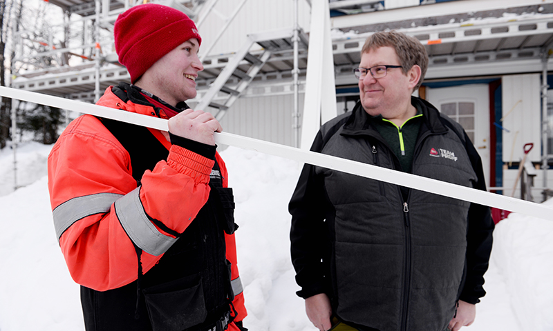 To arbeidere i rødt og svart vintertøy holder hvit planke foran snødekt stillas.