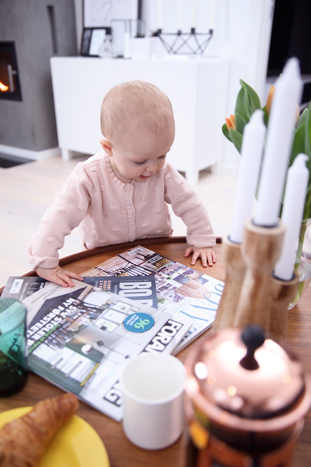 Baby i rosa genser ved rundt trebord med magasiner, hvit kopp, croissant og kobber presskanne