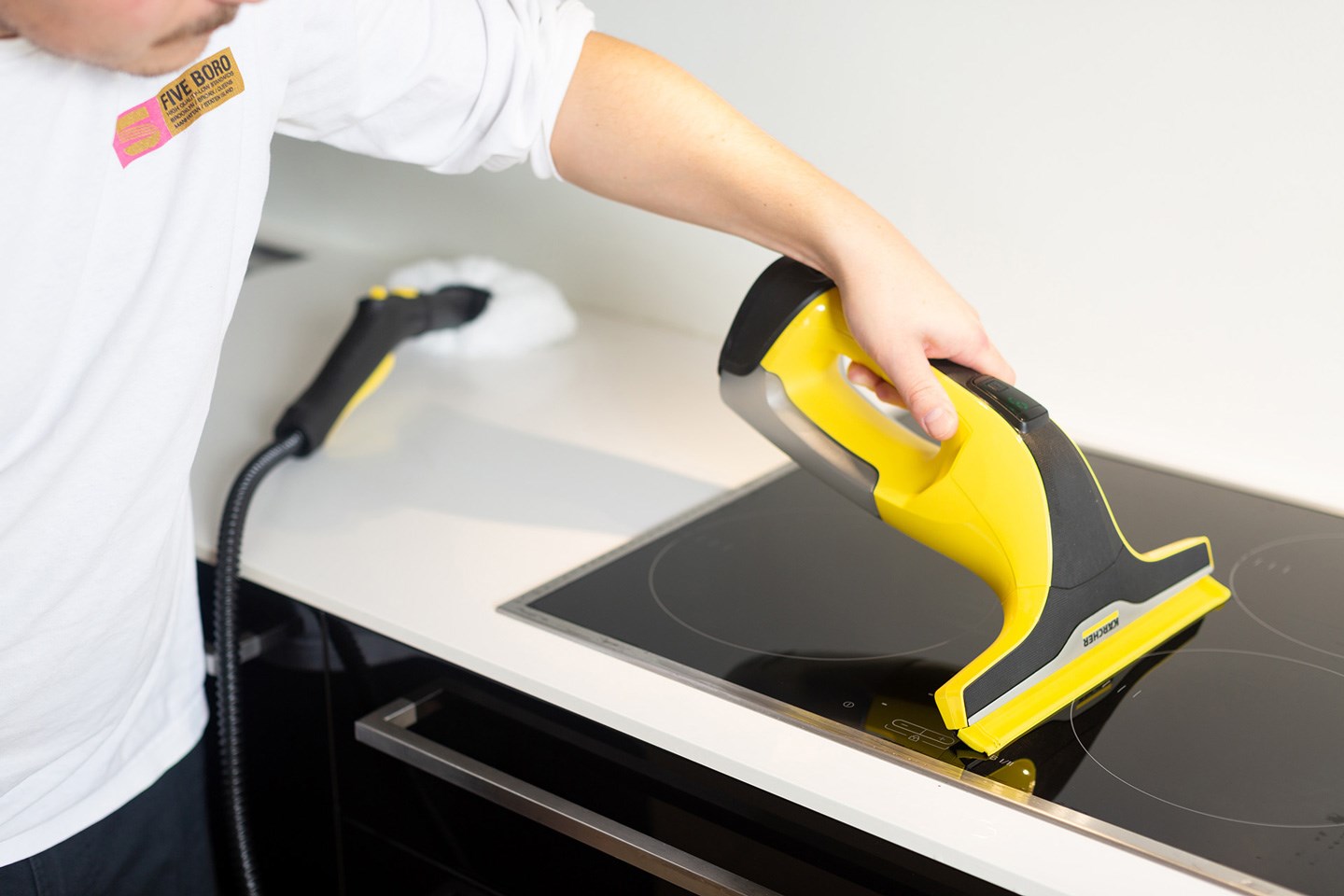 Person rengjør svart keramisk komfyrtopp med gult og svart rengjøringsverktøy.