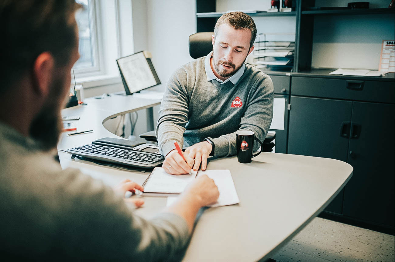 To i grå gensere ved kontorbord, skriver med rød penn, tastatur og svart kaffekopp.
