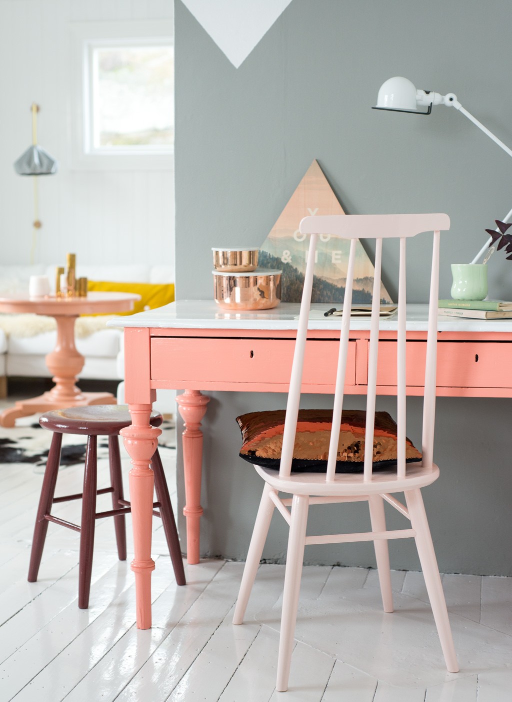 Hjemmekontor med korallrosa skrivebord, hvit trestol med kobberpute og kobberbokser mot grå vegg