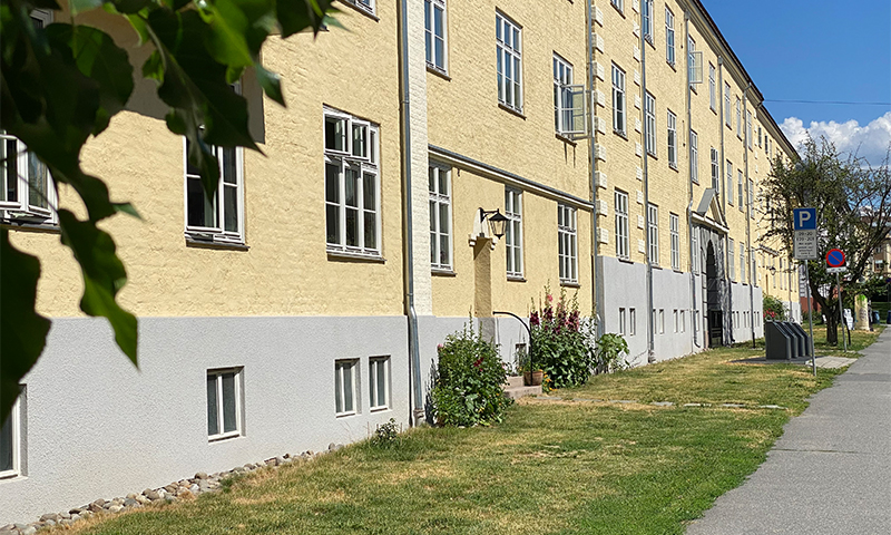 Langt gult boligbygg med grå sokkel, vinduer, blomsterbed foran grønn plen under blå himmel.