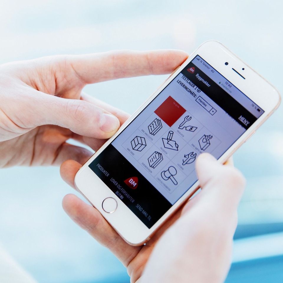 Hender holder hvit mobil med Byggmakker-app: ni ikoner i sort, hvitt og rødt på lys bakgrunn.