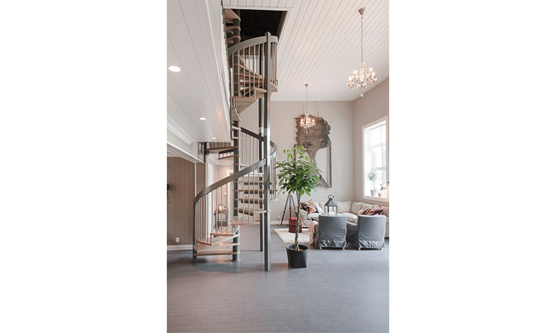 Lyst oppholdsrom med grå spiraltrapp, hvite møbler, grønn potteplante og lysekrone.