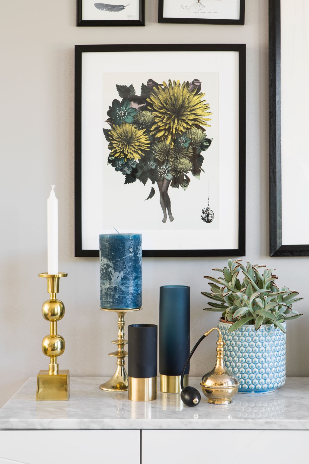 Hvit marmortopp med gulllysestaker, blå lys, sukkulent i blå krukke under plakat med gule blomster