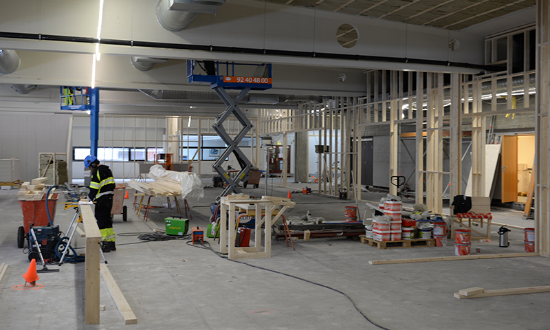 Store industrihall under oppussing med trestendere, blå sakseheis, arbeider i gul-svart drakt