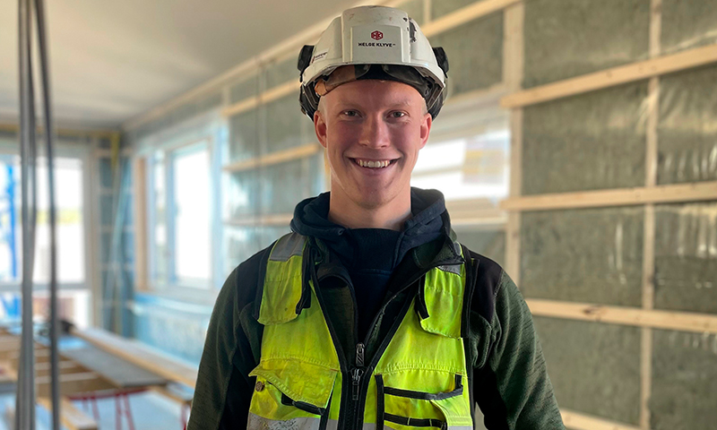 Bygningsarbeider i gul refleksvest og hvit hjelm i isolert råbygg med vinduer og trebjelker