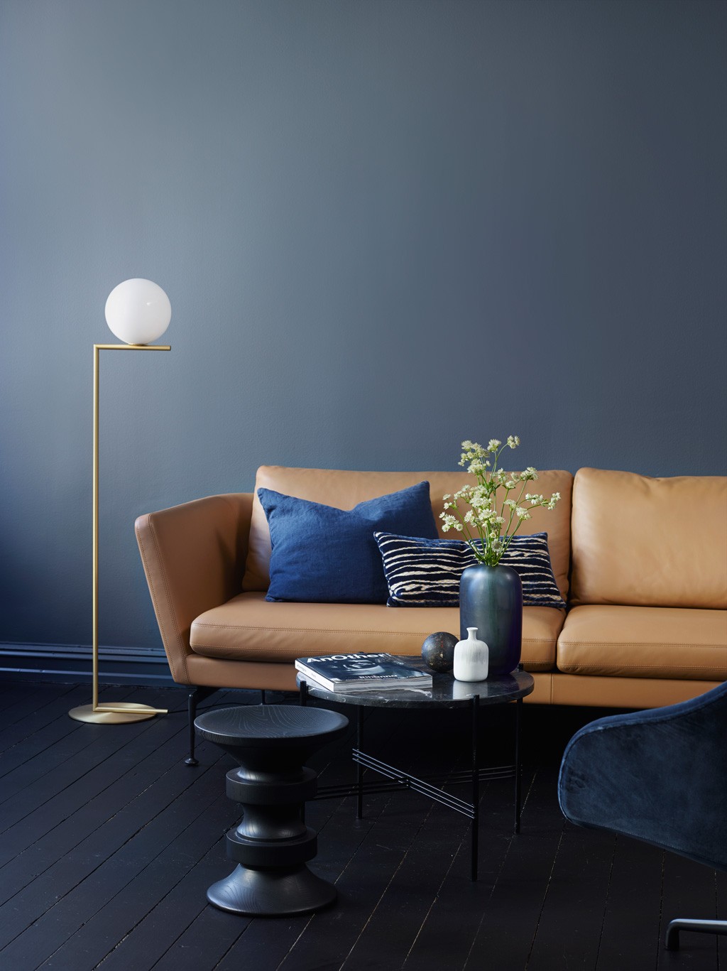 Karamellfarget skinnsofa mot mørkeblå vegg, gullgul gulvlampe, sorte bord med vase og hvite blomster.