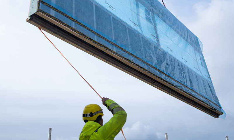 Byggearbeider i gul vernedrakt og hjelm holder tau mens et blått glasspanel løftes mot skyet himmel