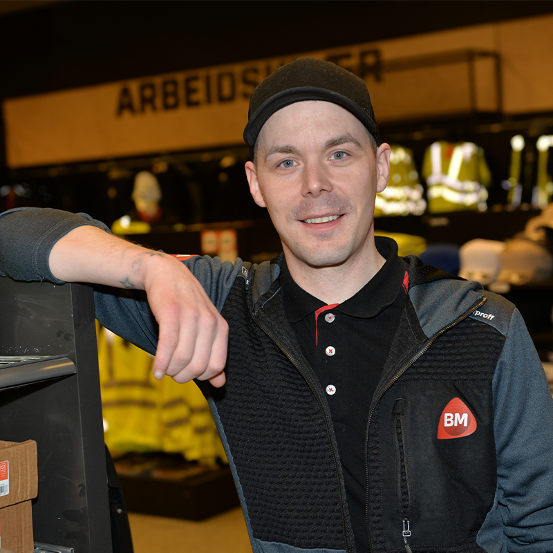 Ansatt i svart BM-genser i byggevarebutikk med verneklær og reflekser i bakgrunnen