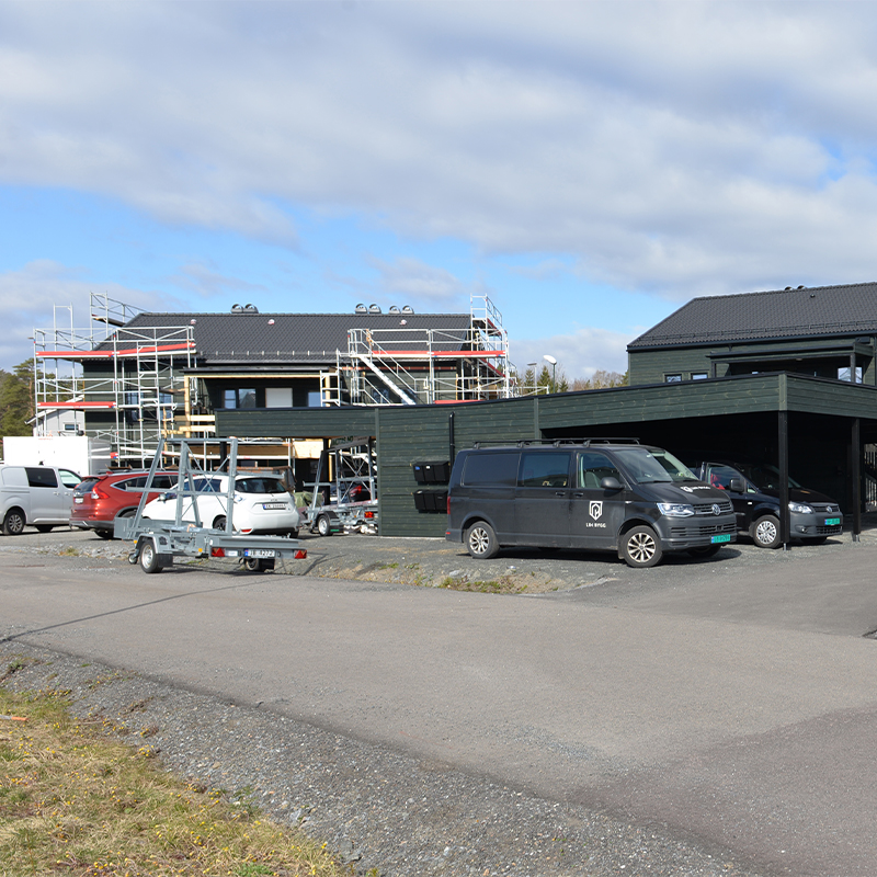 Mørkt trehus under oppussing med stillas, flere parkerte biler og grå himmel.