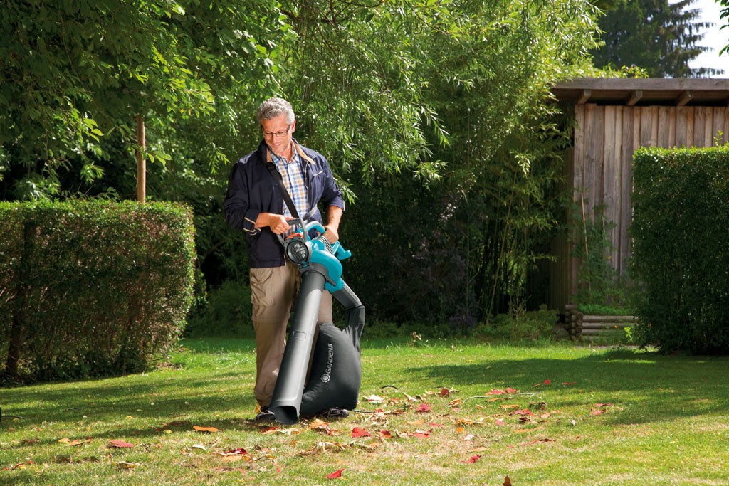 Mann i hagetøy rydder løv med blå løvblåser på grønt gress foran hekk og trær.