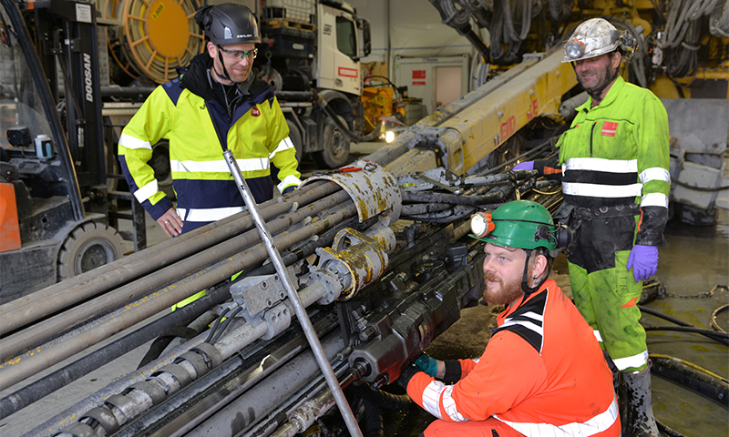 Tre arbeidere i gule, grønne og oransje verneklær ved stor borerigg inne på anleggsverksted