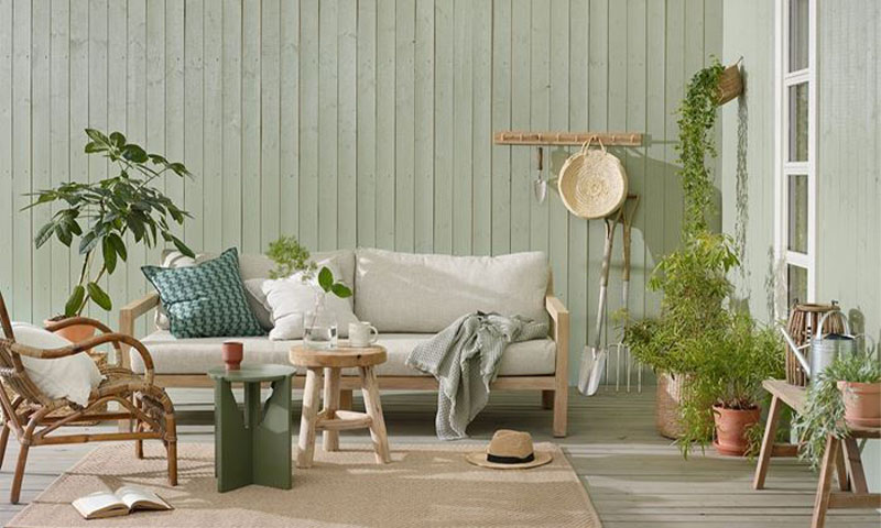 Utemøbelområde med lysegrønn panelvegg, kremhvit sofa, rattanstol, trebord og grønne planter