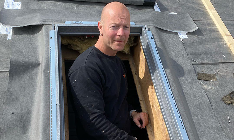 Person i svart genser kikker ut av takvinduåpning med grå metallramme på svart takbelegg.