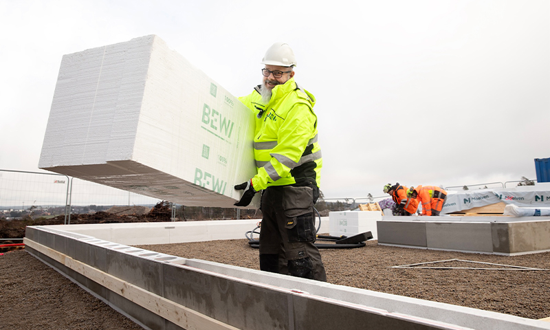Byggearbeider i gul jakke og hvit hjelm løfter stor hvit BEWI-blokk på fundament under grå himmel