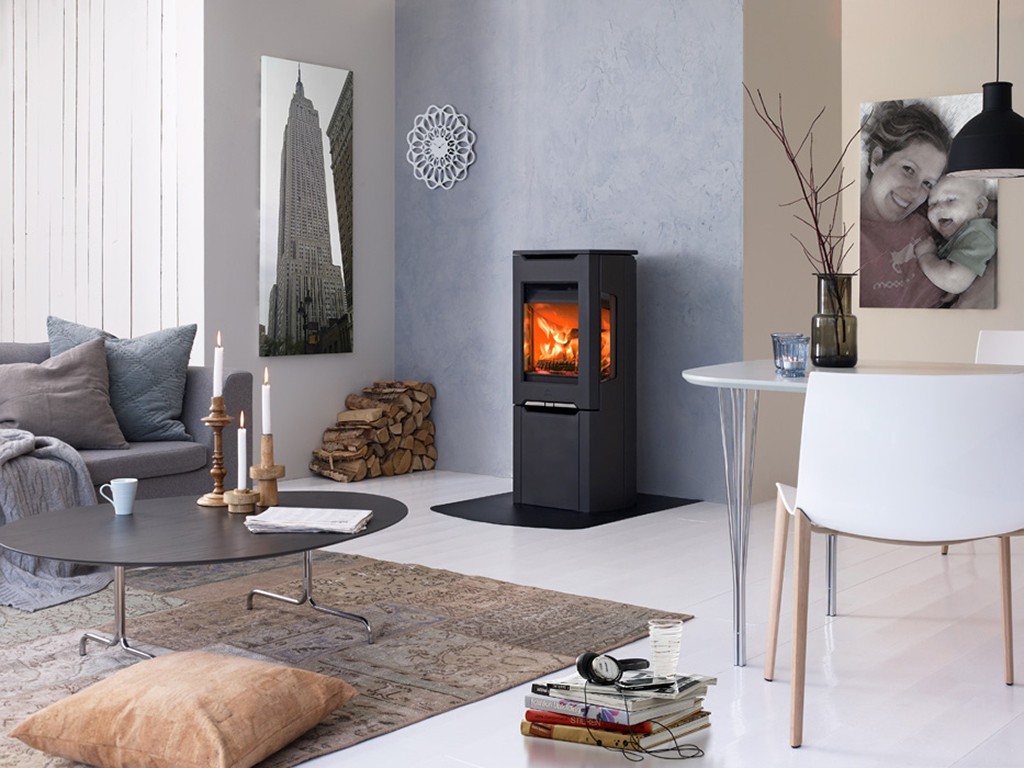 Moderne stue med grå sofa, svart vedovn med flammer, vedstabel, rundt svart bord, hvite fliser