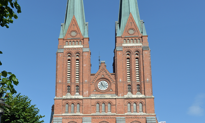 Rød teglsteinskirke med to grønne spir og sentralt klokketårn mot klar blå himmel