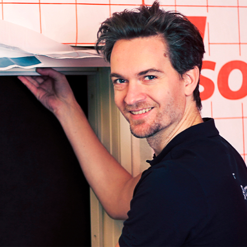 Mann i mørk skjorte monterer hvitt byggeunderlag med rødt rutenett over døråpning.