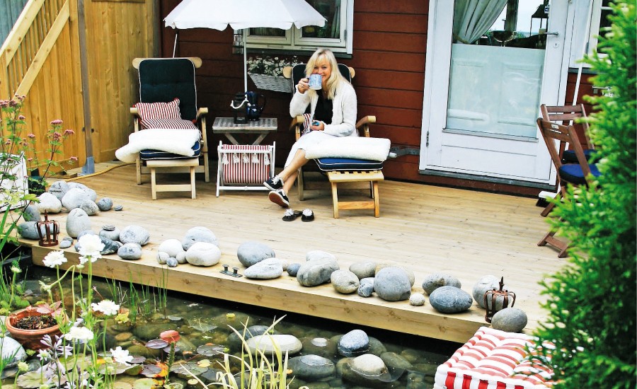 Kvinne på tredekke ved rødt hus med hvit parasoll, tekopp i hånd, steiner langs dam med vannliljer.