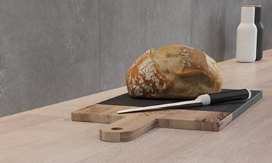 Gylent brød på lysebrun skjærefjøl med svart brødkniv, hvit og svart vase mot grå vegg