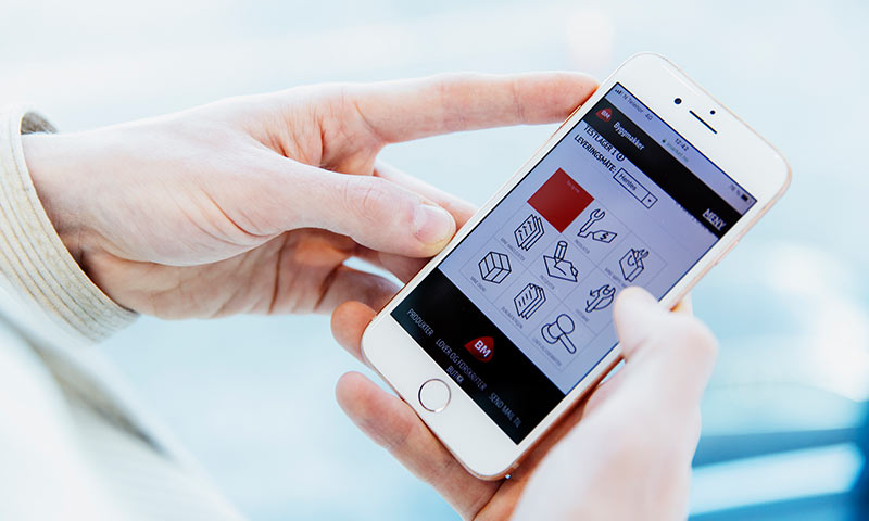 To hender holder hvit mobil med byggerelatert app med røde og blå ikoner på lys bakgrunn