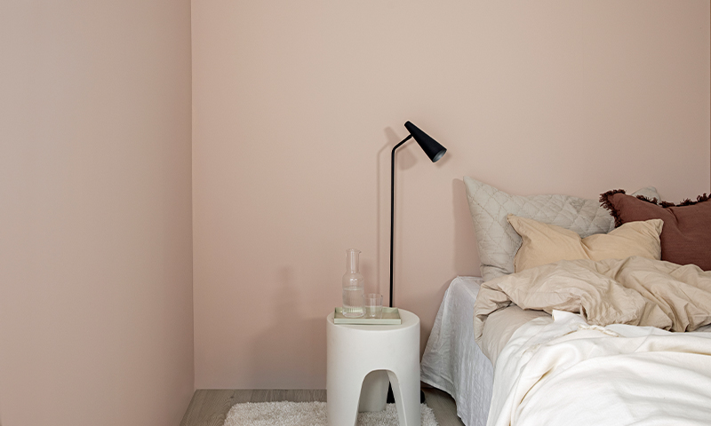 Soveromshjørne: rosa vegg, seng med kremputer, hvitt nattbord med karaffel og svart gulvlampe.