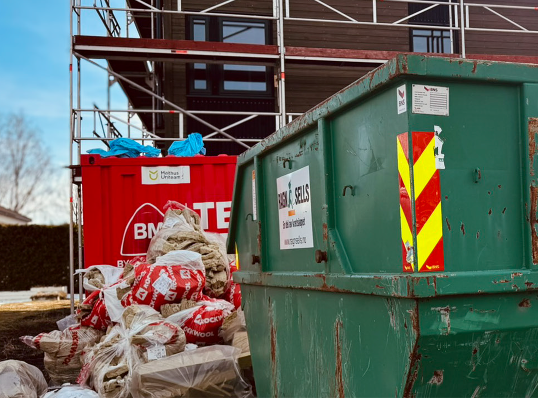 Grønn og rød avfallscontainer på byggeplass med sekker av Rockwool-isolasjon og stillas foran en bygning.