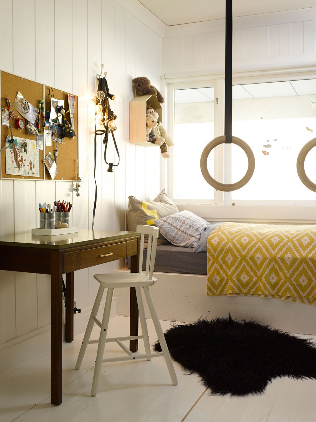 Hvitt barnerom med skrivebord og stol, gul-hvitt pledd på seng, gymringer og kosedyr.