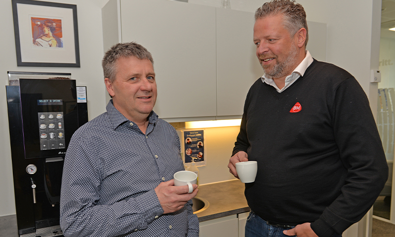 To kolleger ved kaffemaskin i kontorkjøkken, holder hvite kopper ved grå benk.