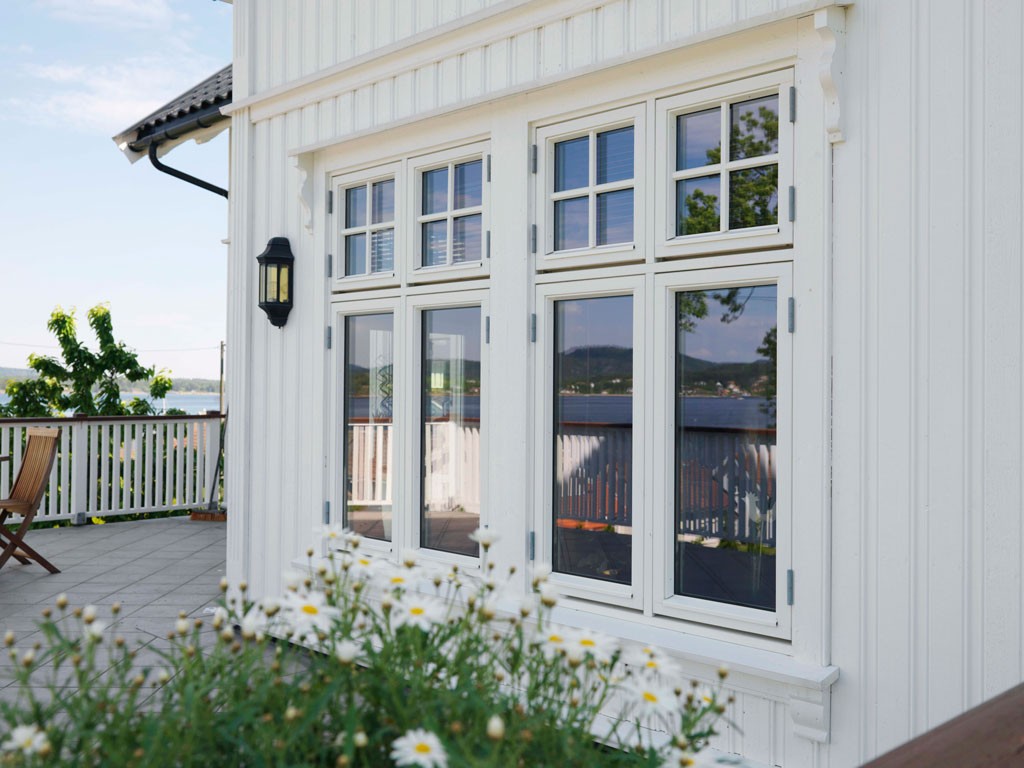Hvit tømmerhus med store vinduer, prestekrager i forgrunnen, terrasse og sjøutsikt.
