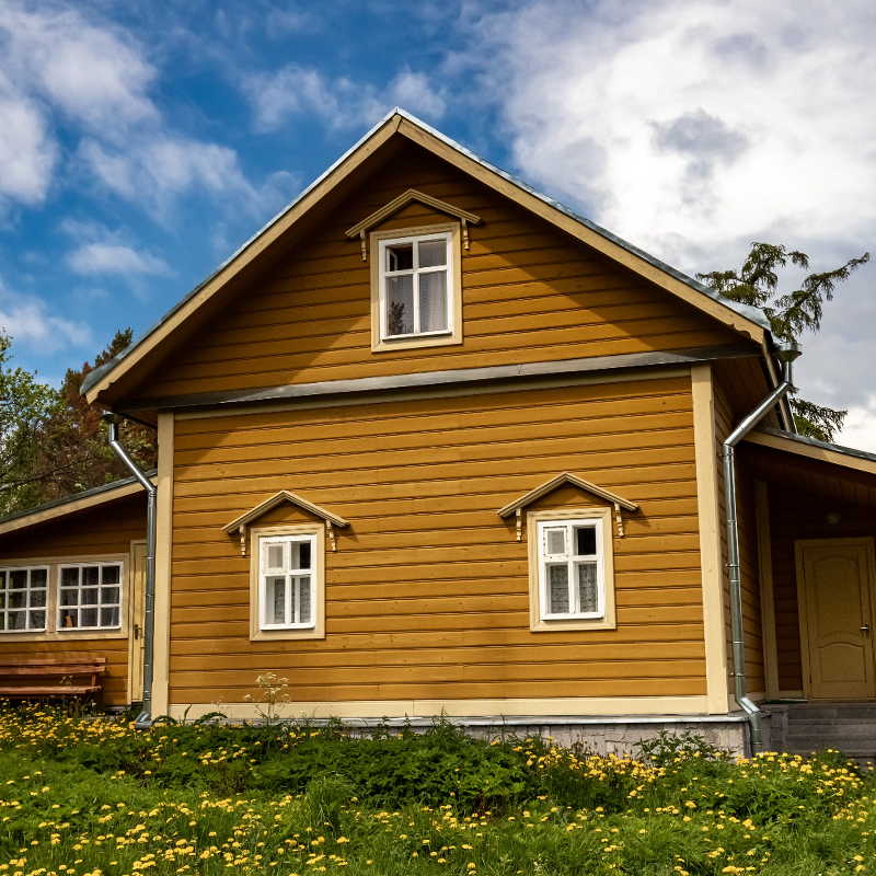 Gult trehus med hvite vindusrammer, gress med gule løvetann og blå himmel.