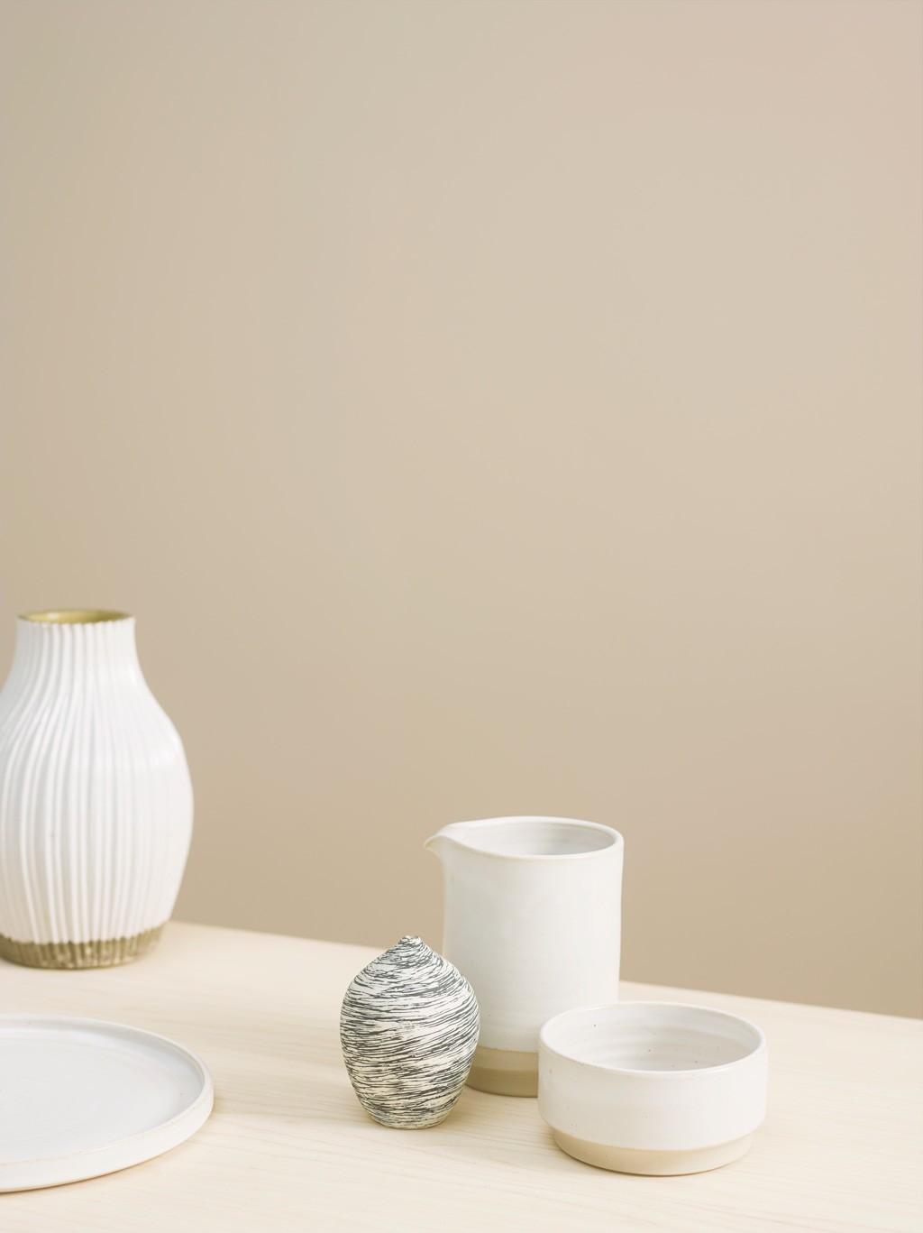 Hvit keramikkvase med gulldetalj, mugge, skål, plate og stripet vase på lyst bord.