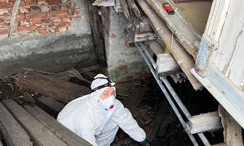 Mann i hvit beskyttelsesdrakt og maske med hodelykt under råttent tregulv