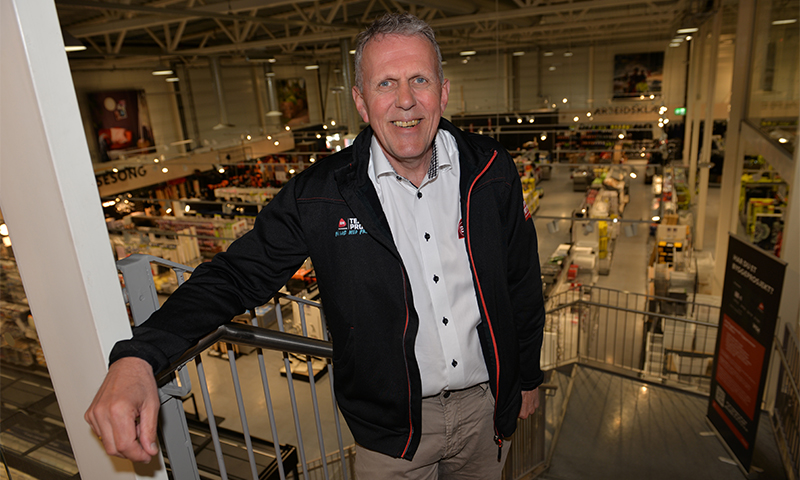 Person i svart jakke og hvit skjorte ved rekkverk i andre etasje i lys byggevarebutikk