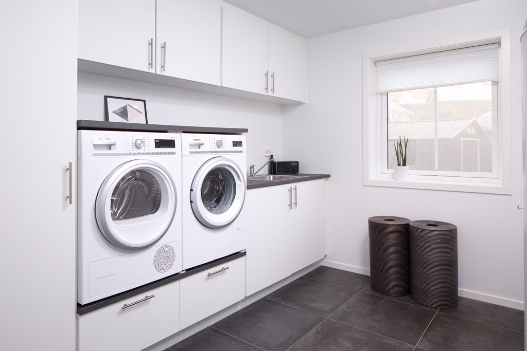Lyst, moderne vaskerom med hvite skap, vaskemaskin, tørketrommel, vask og to mørke kurver