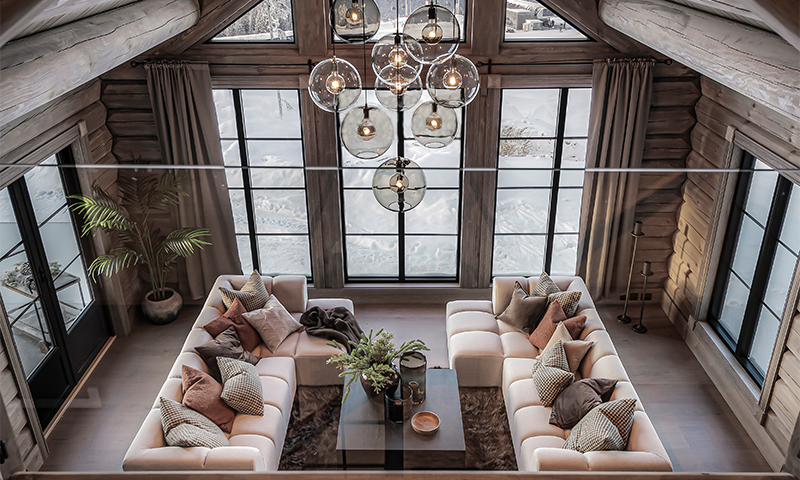 Lyst oppholdsrom i tømmerhytte med to hvite sofaer, glasskulelampe, trebord og snødekt utsikt.