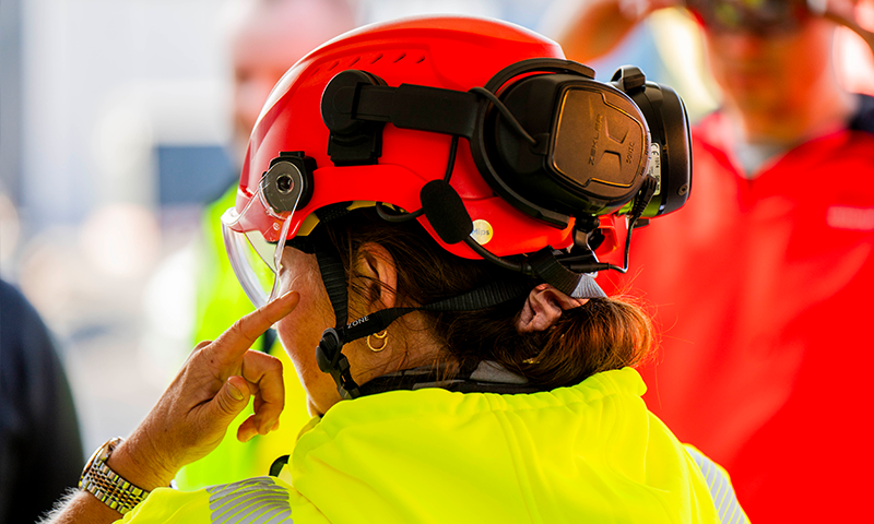 Kvinne i gul vernejakke med rød hjelm og sorte hørselsvern på byggeplass.