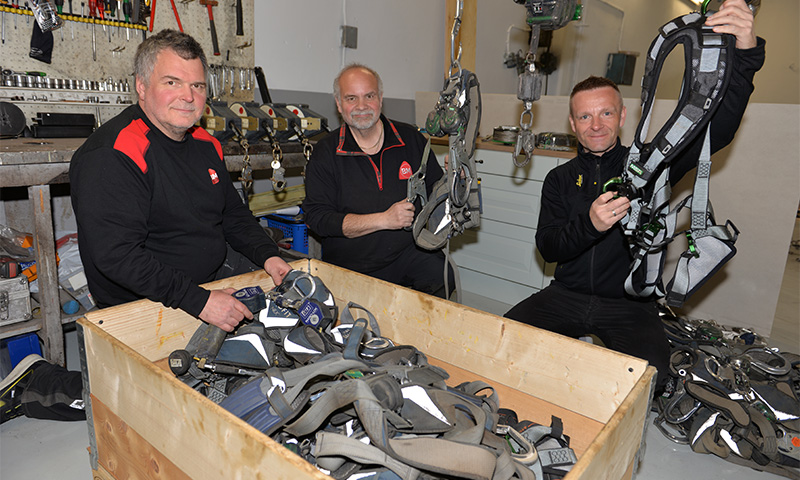 Tre menn i mørke arbeidsklær inspiserer grå-grønne sikkerhetsseler over en trecontainer i verksted