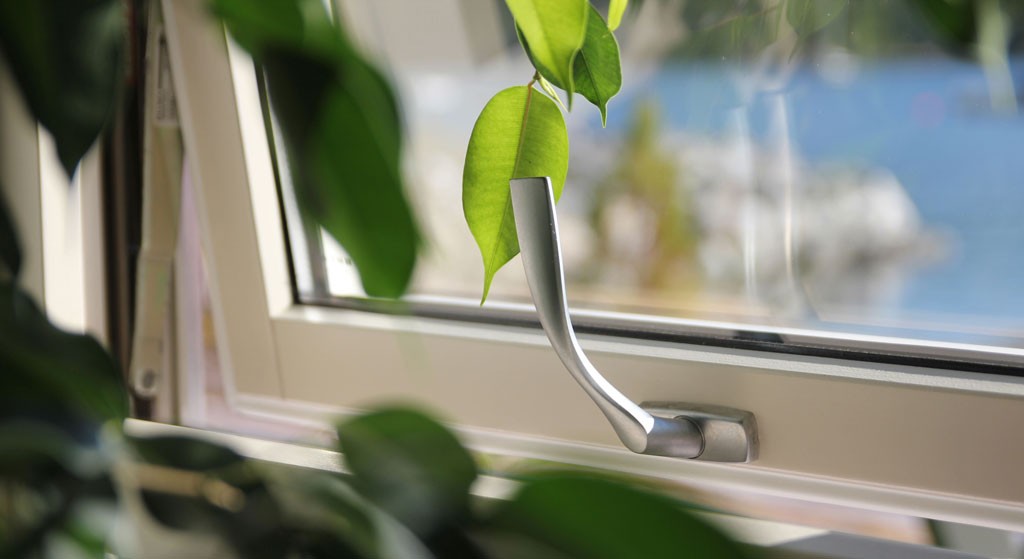 Nærbilde av sølv vindushåndtak på hvit ramme med grønne blader og blå sjø i bakgrunnen