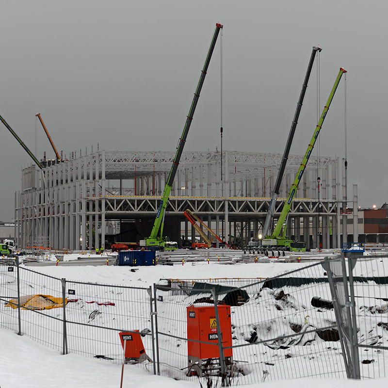 Stålkonstruksjon av bygg under oppføring på snødekt tomt, med grønne kraner.