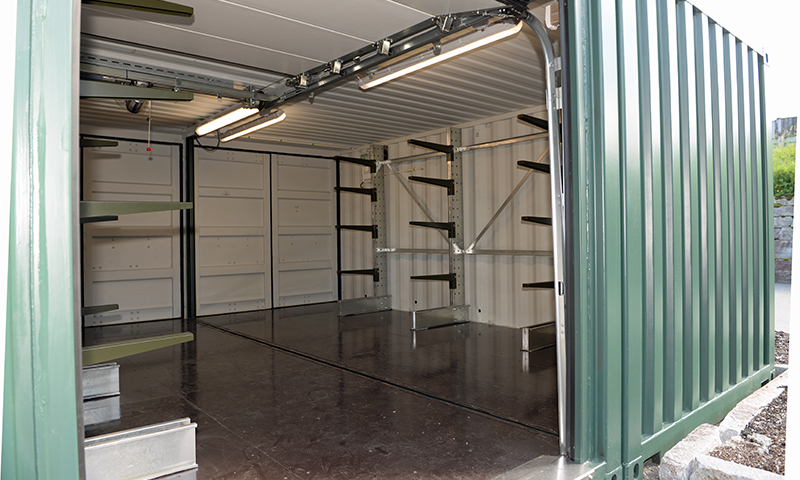 Åpen grønn metallcontainer med tomt, mørkt gulv, hyller og lysrør i taket.