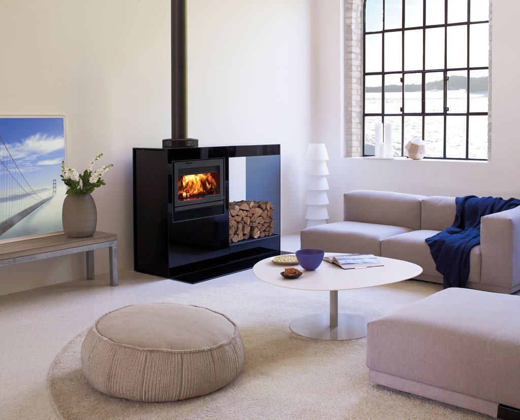 Lyst stue med grå sofa, svart vedovn med ved og ild, hvitt bord og havutsikt