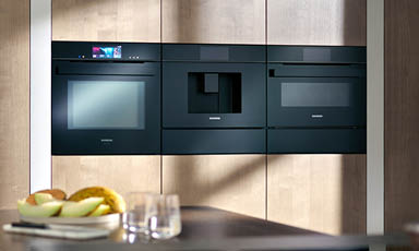 Svart integrert ovn, mikrobølgeovn og kaffemaskin i lyst trepanel, fruktskål på benk.