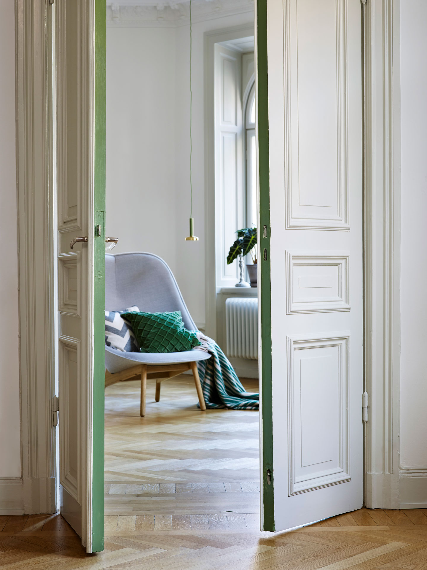 Åpne hvite dører mot rom med grå lenestol på lys parkett, grønne puter, hengelampe og plante