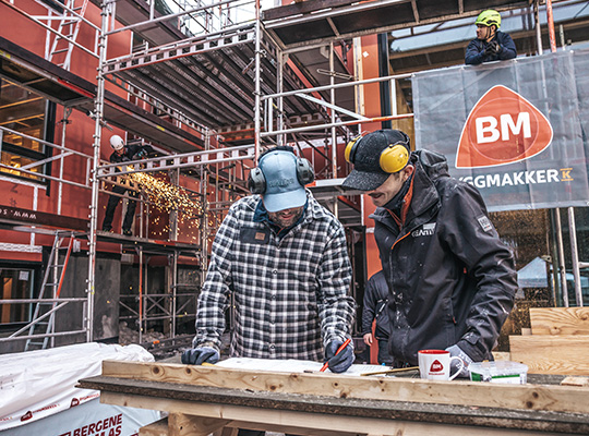 To håndverkere i hjelm og hørselsvern markerer trestokk på byggeplass med røde stålrør og BM-banner.