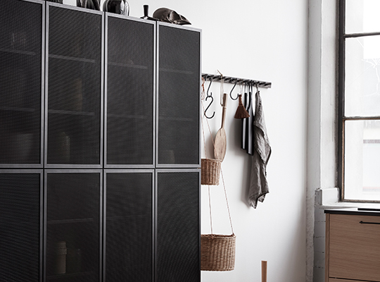 Svart stålgitterskap ved hvit vegg med hengende kurver, jakke og vindu med trekarm.