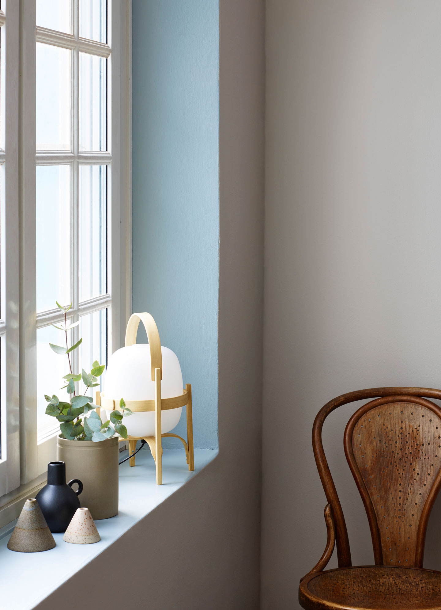 Lyst vindusbord med keramikkvaser, eukalyptus, hvit lampe på lyseblå karm og brun trestol.